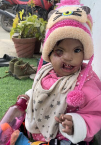 Santoshi, a toddler wears a pink outfit, she sits outside on grass and looks at the camera.