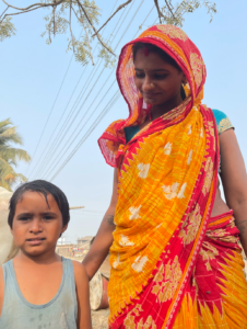 A woman in a bright saree stands looking down at a child with her hand on their shoulder. The child is looking at the camera.