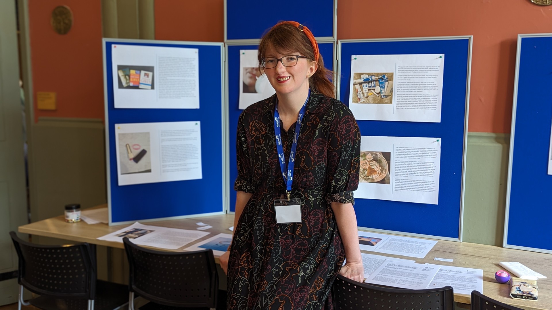 Danielle McWilliams, a white woman with reddish brown hair tied back, a fringe and red headband, a cleft lip, wears a black dress with colourful outlines of faces all over it. She leans back on a table. Behind her is a big blue board with print outs of her research on it. 