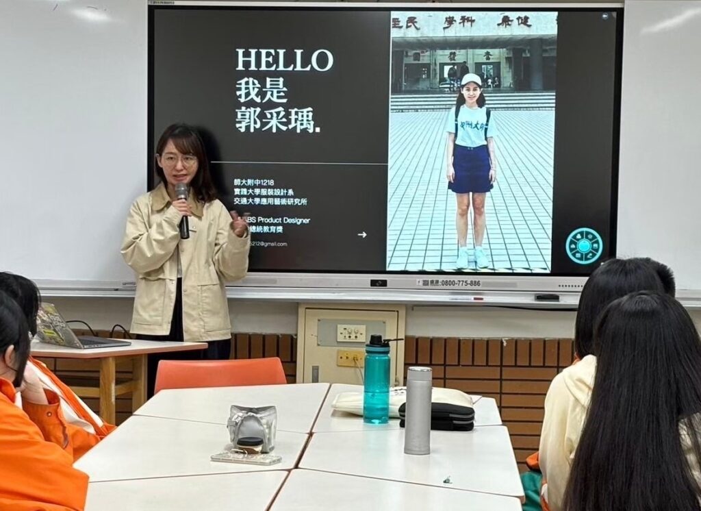 Tsai-Yu stands in front of a presentation screen with a photo of her younger self on it and text, Hello' followed by further text in Chinese. The backs of a group of people sitting around a table look toward Tsai-Yu