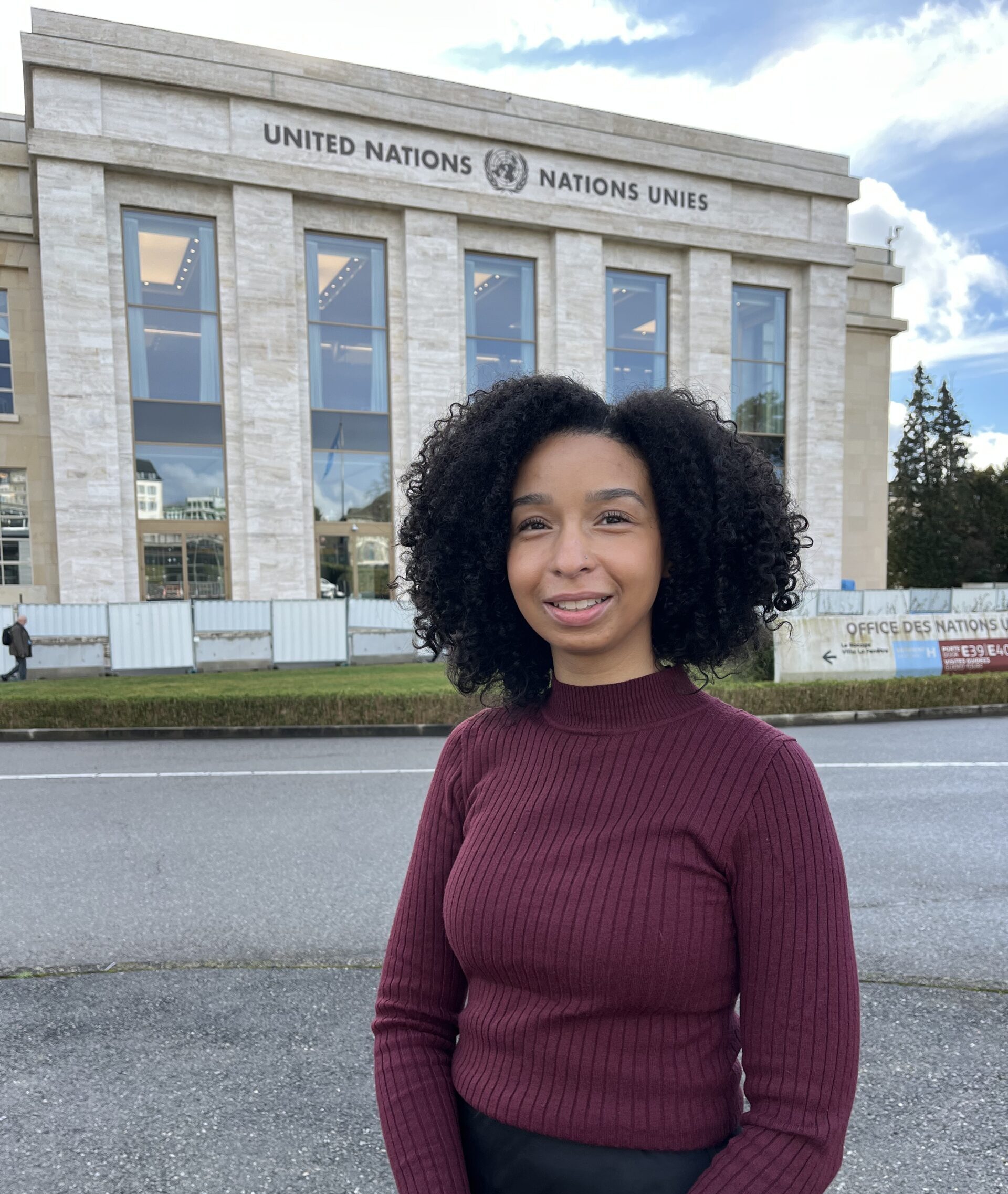 Photo of Chloe who is looking at the camera and smiling, she is wearing a burgundy jumper and is standing in front of the UN Geneva building.