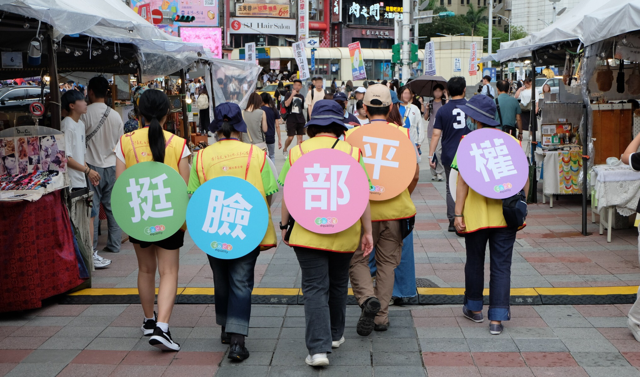 Photo of a busy street in Ximending, Taipei. Five Sunshine volunteers are walking around with on their backs in Chinese letters meaning "Support Face Equality"