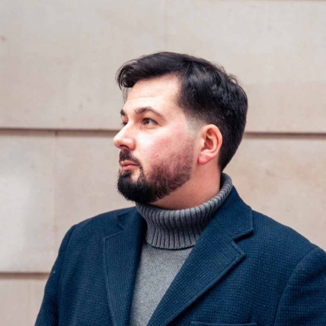 Side-on photo of Dean, a white man with dark short hair and a beard. He is wearing a grey turtleneck jumper, a navy jacket, and is standing against a stone wall.