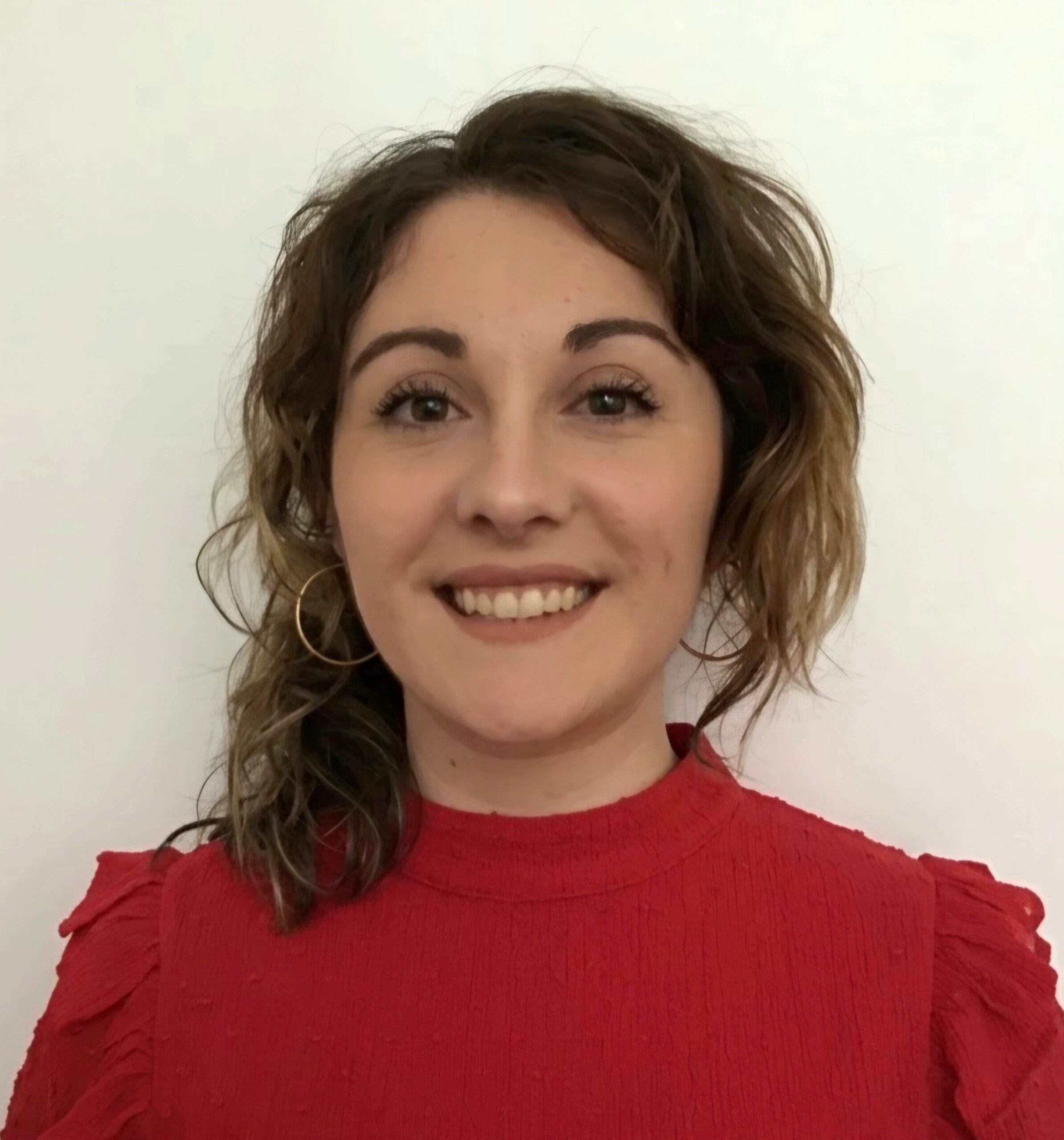 Photo of Rae who is wearing a red top. Rae has dark wavy hair and is smiling at the camera.